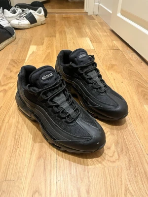 Nike Air Max 95 Triple Black - Säljer ett par Nike Air Max 95 i helsvart färg. Skorna har en chunky sula med synliga Air-enheter, mesh och syntetpaneler samt klassisk snörning. Perfekta för dig som vill ha en clean och sportig look med streetkänsla.