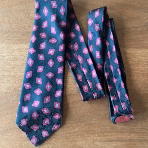 Marinblå elegant slips  - Elegant mörkblå slips med rosa och vita geometriska mönster. Slipsen har en diskret paisleystruktur i tyget som ger extra detalj. Perfekt för dig som vill sticka ut lite extra med accessoarerna. Materialet är silke och slipsen är oanvänd. 