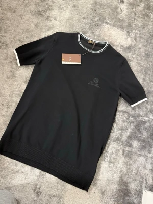 Svart t-shirt från Loro Piana - Svart t-shirt från Loro Piana med broderad logga på bröstet. T-shirten har vita detaljer vid krage och ärmslut samt ribbad nederkant. Klassisk och stilren modell i mjuk bomull, perfekt för en clean look.