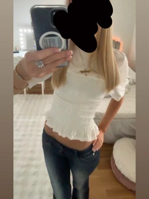 Vit smockad blus med volangkant - Supersöt vit blus med smockad midja och volangkant nertill. Toppen har korta puffärmar och är croppad, vilket ger en fräsch och trendig look. Perfekt att matcha med jeans eller kjol för en stilren outfit.