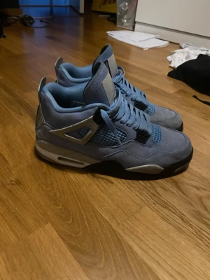 Nike Air Jordan 4 University Blue - Säljer ett par Nike Air Jordan 4 University Blue i storlek 44. Skorna har en snygg ljusblå mocka-överdel, grå detaljer med stänk, svart och vit sula samt meshpaneler. Klassisk Jumpman-logga på plösen och nummer 23-detalj. Perfekt för dig som vill sticka ut med dina sneakers.