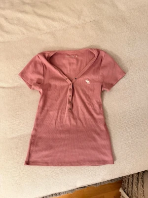 Rosa topp från Abercrombie & Fitch - Säljer en ljusrosa ribbad topp från Abercrombie & Fitch med v-ringning och knappdetaljer framtill. Storlek Xs 💖