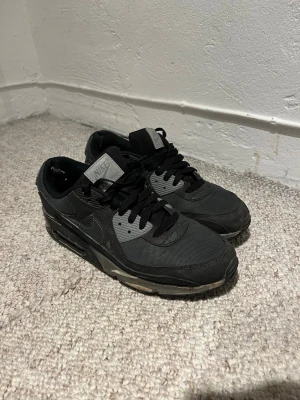 Svarta Nike Air Max sneakers - Säljer ett par svarta Nike Air Max sneakers med klassisk siluett och synlig luftdämpning i sulan. Skorna har mesh och syntetpaneler, svarta snören och grå detaljer vid snörningen och på plösen. Perfekta för dig som gillar streetwear och sportig stil.