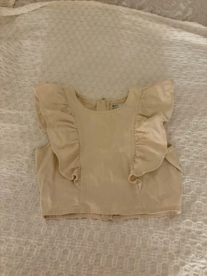 Beige croppad topp med volang - Säljer en beige croppad topp från Gina Tricot med breda volangdetaljer vid axlarna och rund halsringning. Toppen har en synlig dragkedja i ryggen och är tillverkad i jeans tyg. Endast använd en gång😍