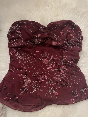 Vinröd paljettprydd tubtopp - Säljer en vinröd tubtopp med glittriga paljetter och små pärlor i blommönster. Toppen är axelbandslös och har en bandeauformad passning som sitter snyggt. Perfekt för dig som vill sticka ut med lite extra bling. Köpt på Vinted står inget märke 