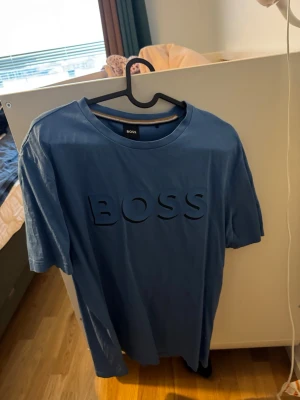 Blå BOSS t-shirt med stor logga - Säljer en blå t-shirt från BOSS med stor, upphöjd logga på bröstet. Klassisk rund hals och korta ärmar. Tillverkad i mjuk bomull som känns skön mot huden. Perfekt för dig som gillar stilrena plagg med tydlig branding.