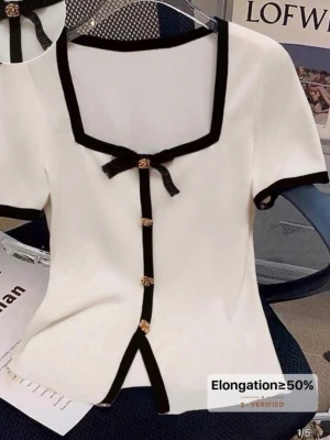 Vit topp med svarta detaljer och rosett - Supersöt vit topp med svarta kanter och dekorativa guldknappar framtill. Fyrkantig ringning, korta ärmar och en svart rosett vid halsen ger en chic vibe. Toppen är lätt ribbad och har en liten slits framtill. Perfekt till jeans eller kjol för en trendig look. Storleken är S/XS