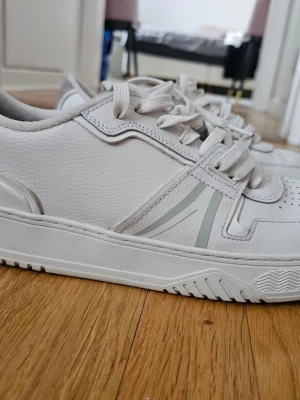 Vita sneakers från Lacoste strl 46 - Säljer ett par fräscha vita sneakers från Lacoste i storlek 46. Skorna har snygga detaljer i grå mocka på hälen, perforeringar på tån och klassisk snörning. Tillverkade i skinn och syntet med diskreta grå ränder på sidan. Perfekta för dig som gillar stilrena och sportiga skor.