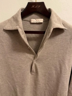 Beige pikétröja från Luca Faloni - Säljer en stilren beige pikétröja från Luca Faloni i 100% bomull. Tröjan har klassisk krage, lång ärm med knappar vid manschetten och en avslappnad passform. Perfekt för dig som gillar en clean och tidlös look.