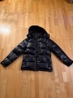 Svart dunjacka med huva från Polo Ralph Lauren - Säljer en svart dunjacka från Polo Ralph Lauren med huva och dragkedja. Jackan har en puffig look, två sidofickor med dragkedja och en liten röd logga på bröstet. Perfekt för kalla dagar och riktigt snygg till streetwear-stilen.