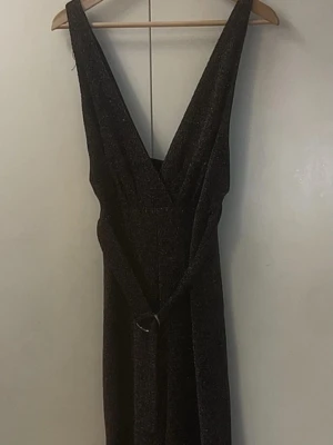 Glittrig svart jumpsuit med bälte - Supersnygg svart jumpsuit med glittrigt tyg och djup v-ringning. Jumpsuiten har ett matchande bälte i midjan och är ärmlös, vilket ger en elegant och trendig look. Perfekt för dig som vill sticka ut med lite extra glitter.