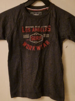 Mörkgrå t-shirt från Produkt, strl 128 - Snygg mörkgrå t-shirt från Produkt med tryck i rött och vitt på bröstet. T-shirten har rund hals och korta ärmar. Tillverkad i ekologisk bomull för skön känsla. Hund finns i hemmet. Kolla in mina andra annonser för samfrakt! 