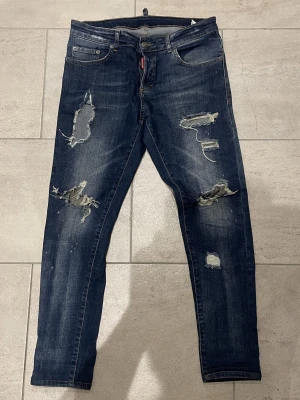 Mörkblå distressed jeans Dsquared2 - Säljer ett par mörkblå jeans från Dsquared2 med slitna detaljer och stora ripped hål på benen. Klassisk femficksmodell med orange sömmar och smal passform. Tillverkade i Italien i mjukt bomullsjeans-tyg. Perfekta för en edgy streetstyle-look.