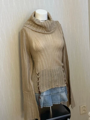 Beige stickad polotröja med snörning - Supersnygg beige stickad polotröja med breda ärmar och cool snörningsdetalj i sidorna. Tröjan har ett mönstrat parti nedtill och vid kragen, samt ribbade partier på ärmarna. Perfekt för dig som gillar unika detaljer och mysig stil.