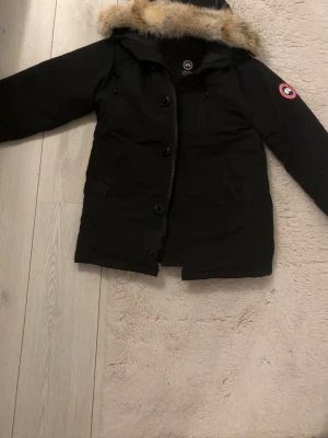  Canada Goose jacka - Säljer en svart dunjacka från Canada Goose med fluffig päls runt huvan och klassisk logga på ärmen. Jackan har dragkedja och knappar framtill, stora fickor och är riktigt varm och skön för kalla dagar. Perfekt för dig som vill ha en ikonisk vinterjacka. Äkta!