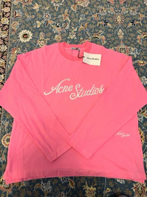 Rosa långärmad tröja Acne Studios - Säljer en rosa långärmad tröja från Acne Studios med vit broderad logga framtill och på ärmen. Tröjan har en klassisk rund halsringning och är gjord i mjuk bomull. Perfekt för dig som gillar stilrena och trendiga plagg. Aldrig använd ny med taggar 
