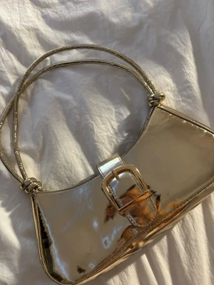 Glansig guldig axelväska med spänne - Säljer en superfin axelväska i glansigt guldigt material. Väskan har en snygg rektangulär form, dubbla axelband och ett stort dekorativt spänne framtill. Perfekt för dig som vill sticka ut med en metallic look och addera lite extra bling till outfiten.