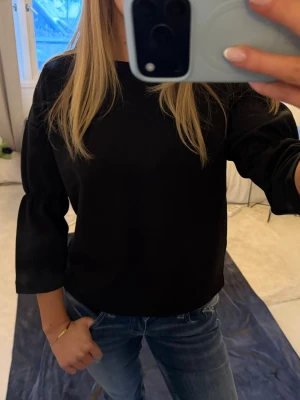 Svart blus från Lindex med vida ärmar - En svart blus från Lindex i storlek S. Modellen har rund halsringning och trekvartsärmar med lätt puff och rynk vid ärmslutet. Tidlös och enkel design som passar till jeans eller kjol.