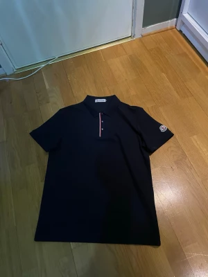 Moncler pikétröja med logga - Säljer en svart pikétröja från Moncler med klassisk krage och knappar framtill. Tröjan har Moncler-logga på ärmen och en snygg detalj med röd och vit rand längs knappslån. Tillverkad i bomull och har korta ärmar.