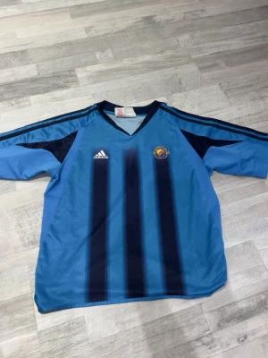 Blå adidas fotbollströja med ränder - Säljer en blå fotbollströja från adidas med mörkblå vertikala ränder och klubbmärke på bröstet. Tröjan har v-ringning, klassiska adidas-ränder på axlarna och är tillverkad i lätt polyester. Perfekt för dig som gillar sportig stil.