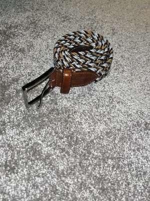 Flätat bälte från Saddler - Säljer ett snyggt flätat bälte från Saddler med detaljer i brunt skinn och metallspänne. Bältet har en mix av blå, beige, vit och bruna toner i ett rutigt mönster. Perfekt accessoar för att lyfta din outfit.