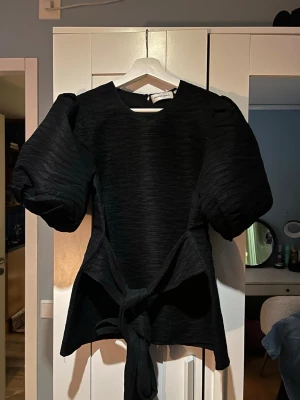 Svart blus med puffärmar och knyt - Säljer en svart blus från Carin Wester med stora puffärmar och snygg knytning i midjan. Blusen har en strukturerad yta. Perfekt för dig som vill ha en statement topp med cool siluett. Använd en gång endast!