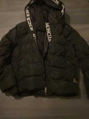 Svart Moncler pufferjacka med huva - Säljer en riktig svart pufferjacka från Moncler i storlek M. Jackan har en stor huva med Moncler-logga längs kanten och dragkedja framtill. Klassisk Moncler-patch på ärmen och coolt serietryck på insidan. Perfekt för dig som vill ha en varm och snygg jacka med streetkänsla.