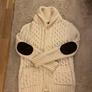 Vit stickad kofta från Divided - Mysig vit stickad kofta från Divided by H&M med grovt kabelstickat mönster, stora fickor fram och svarta armbågslappar. 