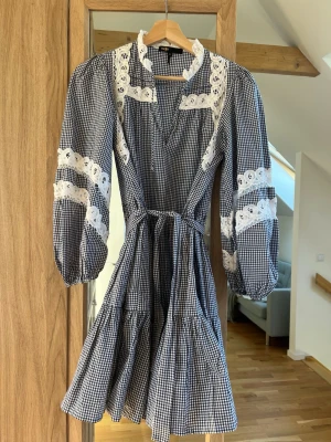 Rutig klänning med spets från Maje - Supersöt kort rutig klänning från Maje med volangkjol, långa puffärmar och vita spetsdetaljer. Klänningen har knytband i midjan och en v-ringad halsringning. 