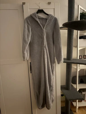 Grå teddy-overall med huva - Supermjuk och mysig grå teddy-overall med huva och långa ärmar. Perfekt för att chilla hemma eller mysa runt i. Overallen är i fluffigt fleece-material och har en relaxed passform.