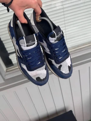 Valentino Rockrunner sneakers blå/vit - Säljer ett par Valentino Rockrunner sneakers i blått, vitt och svart med coolt kamouflagemönster. Skorna har snörning, rund tå och detaljer i mocka och mesh. Perfekta för dig som vill ha något unikt och trendigt på fötterna.