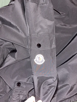 Svart vindjacka från Moncler - Säljer en svart vindjacka från Moncler med huva, dragkedja framtill och två sidofickor med dragkedja. Jackan har en ficka med knapp på ärmen och Moncler-logga. Materialet är lätt och känns sportigt, perfekt för blåsiga dagar.