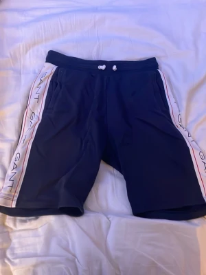 Marinblå shorts från Gant med snörning - Säljer ett par marinblå shorts från Gant med vita sidor och röda detaljer. Shortsen har snörning i midjan och vita sidopaneler med GANT-logga längs benen. Perfekta för chill dagar eller träning. Materialet känns mjukt och sportigt.