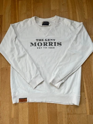 Vit sweatshirt från Morris Division - Vit sweatshirt från Morris Division med svart tryck på bröstet där det står 'THE GENT MORRIS EST TM 1968'. Tröjan har rund hals, långa ärmar och ribbade muddar. Perfekt för en clean och stilren look.