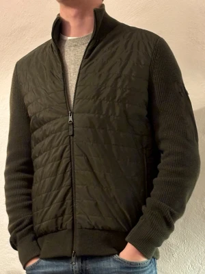 J.Lindeberg Cardigan - Säljer en olivgrön cardigan från J.Lindeberg med quiltad front och stickade ärmar och rygg. Den har dragkedja framtill, hög krage och en liten ficka med logga på ena ärmen. Perfekt för dig som gillar stilren och sportig look. Ny pris 3000kr vårt pris 1699💳, storlek S. Jackan är eftertraktad (säljs ej längre), Ny skick ✅.                                 Hör av dig vid funderingar 📦