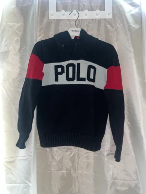 Ralph Lauren tröja  - Stickad tröja från Polo Ralph Lauren i svart med breda vita och röda ränder samt stor POLO-logga framtill. Tröjan har hög krage med knappdetalj och ribbade muddar. Axelbredd 38cm, längd 56cm, armlängd 55cm. OBS plagget är i barnstorlek L (14-16år) men jag skulle bedöma den till XS - men utgå från måtten.  Ifall det inte finns någon bild i annonsen där jag bär plaggen jag säljer så kolla måtten för storlek och googla på modellnamnet för passform 😉 1,19