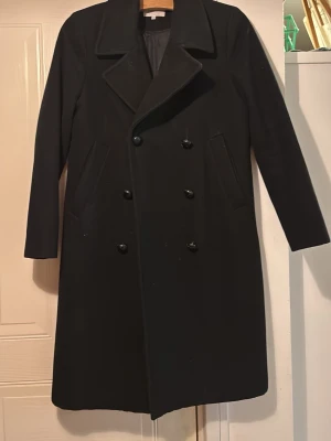 Svart kappa från Second Female  - Stilren svart kappa från Second Female i storlek S. Klassisk dubbelknäppt design med bred krage och raka fickor. Lång modell med helsvart look, perfekt för dig som gillar en clean och tidlös stil. S/38