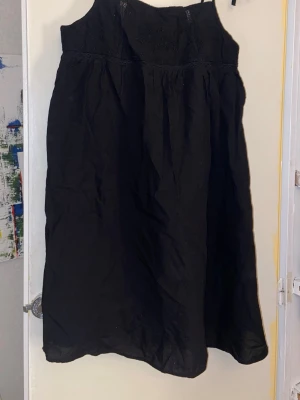 Svart långklänning med broderi - Säljer en svart långklänning med broderade blommor upptill och spetsdetaljer. Klänningen har en luftig passform och är ärmlös, perfekt för varma dagar. Materialet känns som bomull och ger en skön känsla mot huden.