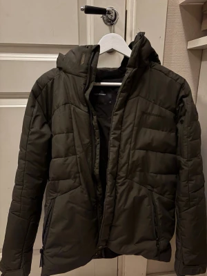 Mörkgrön pufferjacka från Peak Performance - Säljer en mörkgrön pufferjacka från Peak Performance med huva och dragkedja. Jackan har två fickor med dragkedja och quiltad design. Perfekt för kyliga dagar och har en snygg, sportig look. Storlek barn 160, men passar mig som bär xs eller s