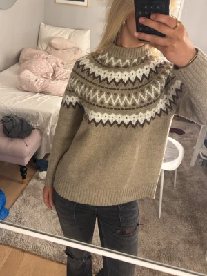 Beige stickad tröja med mönster - Säljer en beige stickad tröja med vitt och brunt zigzag-mönster runt oket. Tröjan har lång ärm, ribbade muddar och en lite högre hals. Använd få gånger.