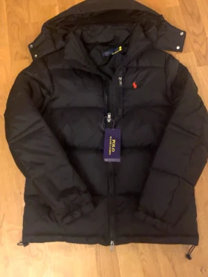 Svart pufferjacka från Polo Ralph Lauren - Säljer en svart pufferjacka från Polo Ralph Lauren med röd broderad logga på bröstet. Jackan har dragkedja framtill, hög krage och huva. Perfekt för kalla dagar och riktigt snygg till streetwear-stilen.