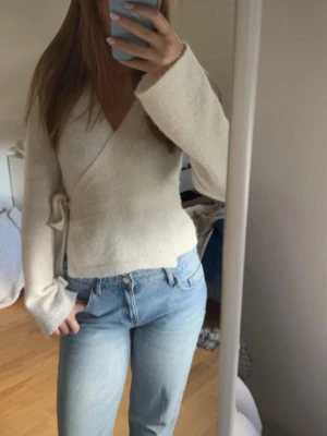 Krämvit omlottkofta med knyt - Mysig krämvit omlottkofta med knyt i sidan och v-ringad hals. Tröjan har långa ärmar och är stickad i ett mjukt material som känns skönt mot huden. Perfekt till jeans för en enkel men snygg look.