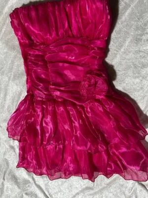 Rosa axelbandslös kortklänning - Säljer en axelbandslös kortklänning i stark rosa färg med glansigt tyg och volanger. Klänningen har en smockad rygg och en dekorativ blomdetalj framtill. Materialet känns som syntet och ger en festlig look med mycket volym och struktur. Storlek 34/36