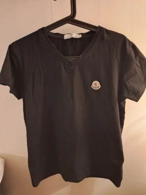 Svart Moncler t-shirt med v-ringning - Svart t-shirt från Moncler i mjuk bomull med klassisk v-ringning och korta ärmar. På bröstet sitter Monclers ikoniska logotypmärke i vitt, rött och blått. Enkel och stilren design som funkar till allt.