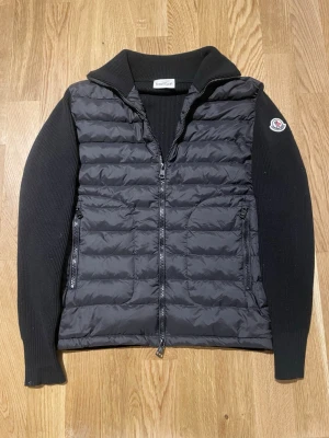 Svart dunväst från Moncler - Säljer en svart dunväst från Moncler med quiltad front och ribbstickade ärmar. Västen har dragkedja framtill, två sidofickor med dragkedja och klassisk Moncler-logga på ärmen. Perfekt för lager-på-lager och snygg till både jeans och hoodies.