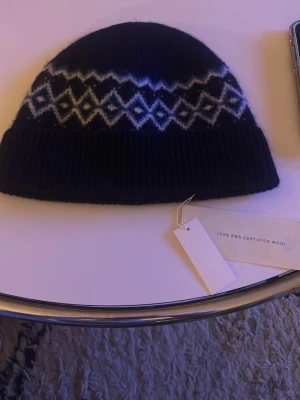 Mörkblå arket mössa 100% ull - Säljer en mörkblå mössa från Arket i 100% RWS-certifierad ull. Mössan har ett snyggt vitt och ljusblått geometriskt mönster runtom och bred ribbad kant. Perfekt för kalla dagar och riktigt skön på huvudet.
