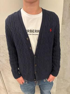 Mörkblå kabelstickad kofta Polo Ralph Lauren - Kabelstickad mörkblå kofta från Polo Ralph Lauren med klassisk röd logga på bröstet. Koftan har v-ringning, knappar framtill och ribbade muddar. Perfekt till jeans och t-shirt för en clean och stilren look. Stoleken är 14-16 år