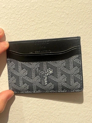 Svart slim plånbok - En Aldrig använd stilren svart kortfodral med grafiskt mönster och flera fack för kort. Tillverkat i skinn med klassisk logga och texten 'Made in France' präglad på ovansidan. Perfekt för dig som vill ha något både snyggt och praktiskt.