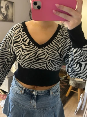 Zebramönstrad croppad stickad tröja - Säljer en croppad stickad tröja med svartvitt zebramönster och v-ringning. Tröjan har långa ärmar och bred svart mudd i midjan och vid ärmsluten. Perfekt för dig som vill sticka ut med ett djurmönster och gillar trendiga detaljer.
