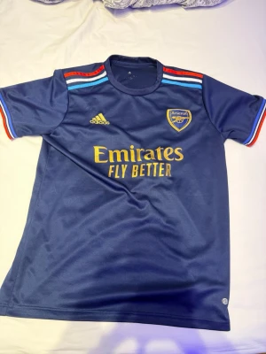 Arsenal bortatröja Saka 7 Adidas - Snygg marinblå Arsenal bortatröja från Adidas med Saka 7 på ryggen. Tröjan har gula detaljer, klubbmärke och Adidas-logga på bröstet samt färgglada ränder på axlar och ärmslut. Tillverkad i polyester, perfekt för fotboll eller att bära till vardags.